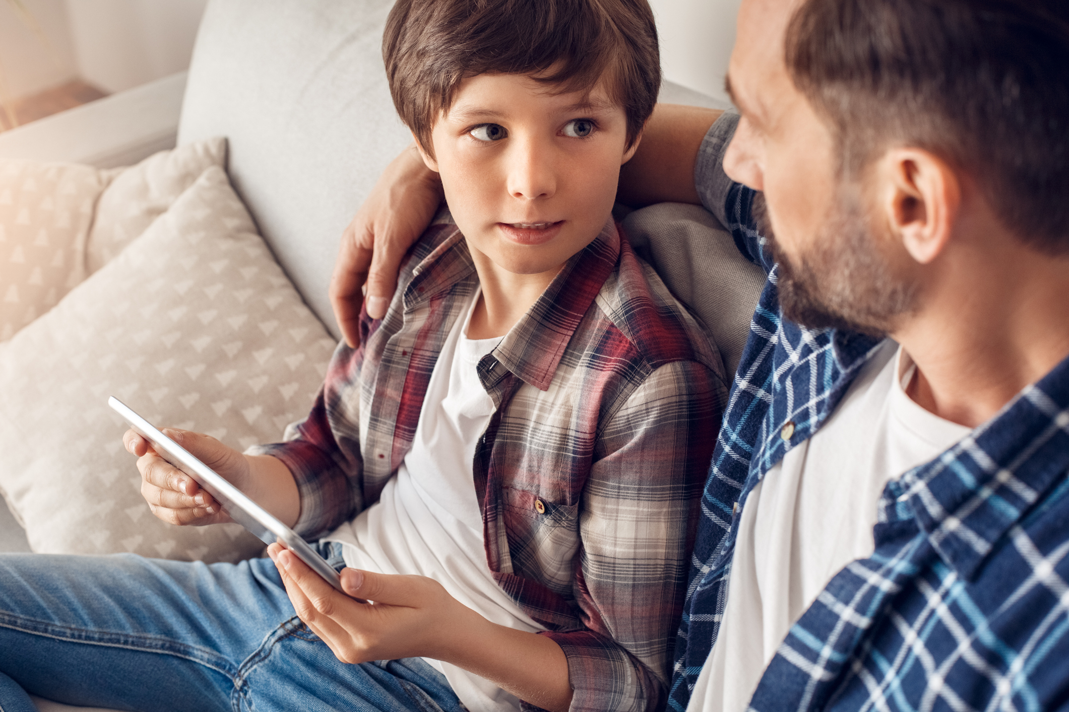 Father and little son together at home sitting on sofa dad hugging boy holding digital tablet looking at each other serious close-up