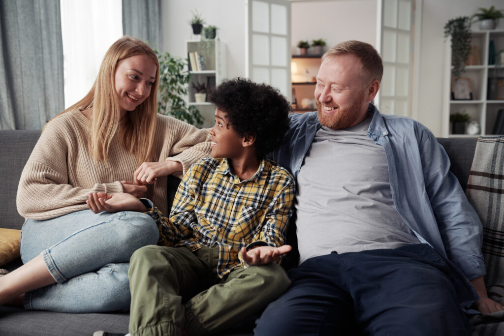 Happy parents talking to their adoptive son African ethnicity while resting on sofa in living room