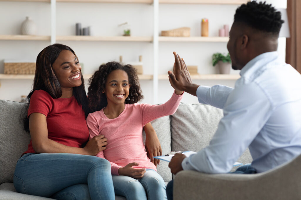 Positive black mother and daughter teenager at the appointment with male psychologist, happy african american girl giving psychotherapist high five, clinic interior, family therapy session concept