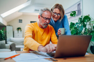 Happy senior couple laughing and bonding while using laptop at home. Smiling elderly husband and wife having fun satisfied with buying insurance, paying bills online. Man showing something on laptop.