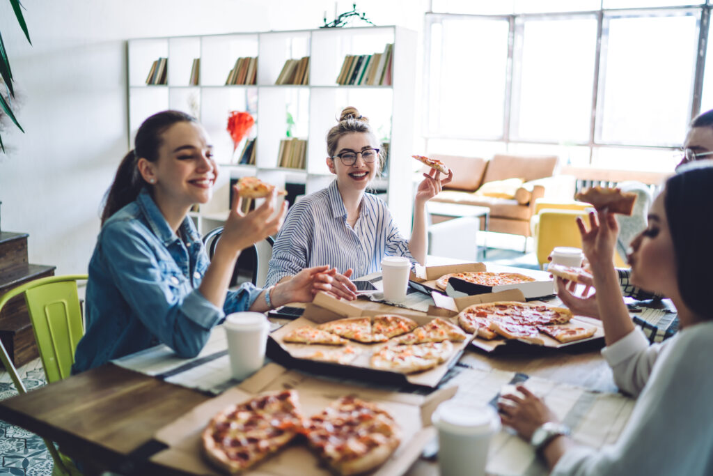 Joyful company of young friends speaking together and laughing at table while eating tasty appetizing pizza in light kitchen