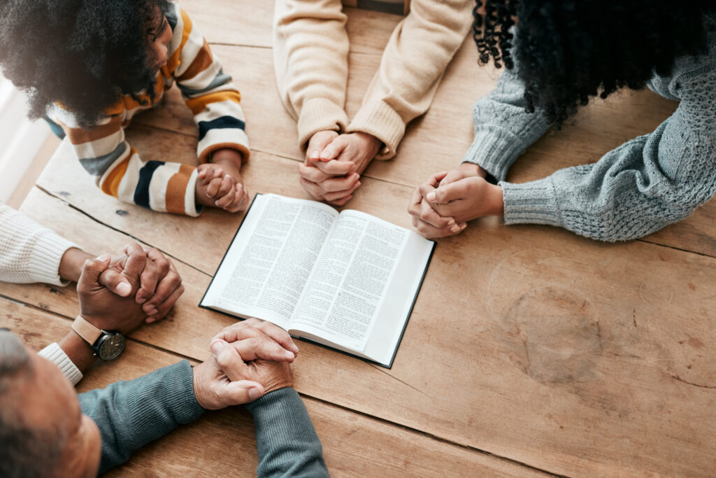 a group of individuals from varying ethnic backgrounds is gathered around a table with bibles in prayer